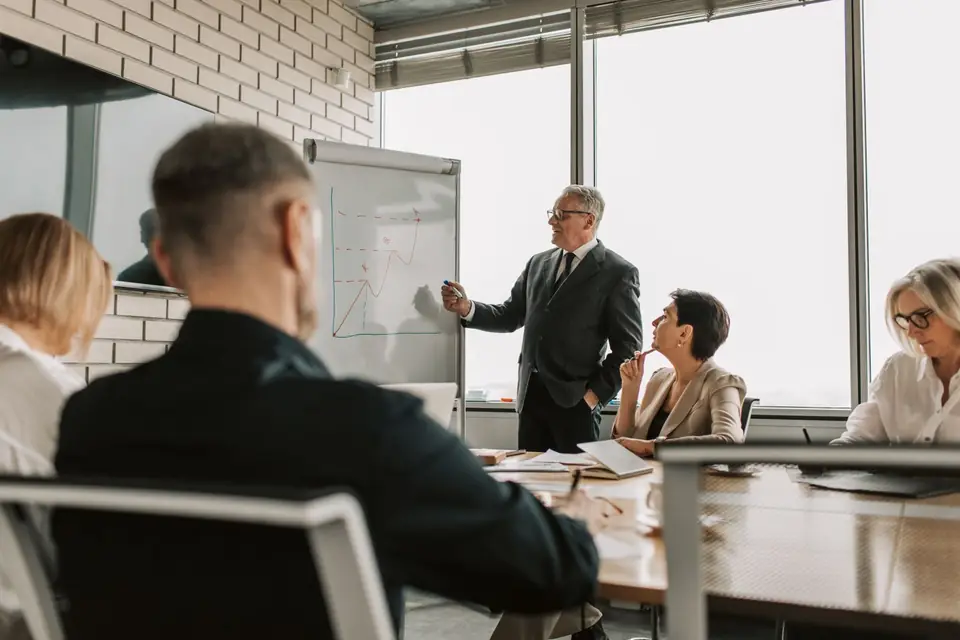 A consultant stands at a flip chart presenting a growth chart to executives seated around a boardroom table.